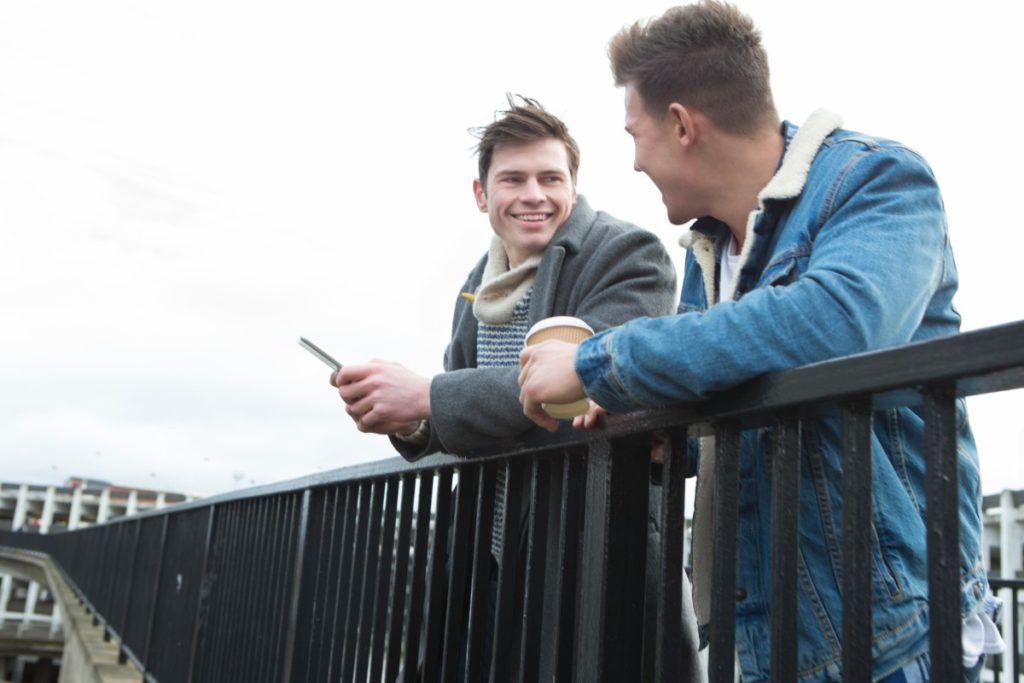 Two young men talking in the city. One is holding a smartphone, the other is holding a disposable coffee cup.